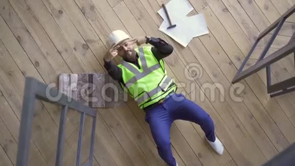 戴着头盔和绿色夹克的快乐微笑的建筑者躺在地板上展示着胜利的姿态抬头视频的预览图