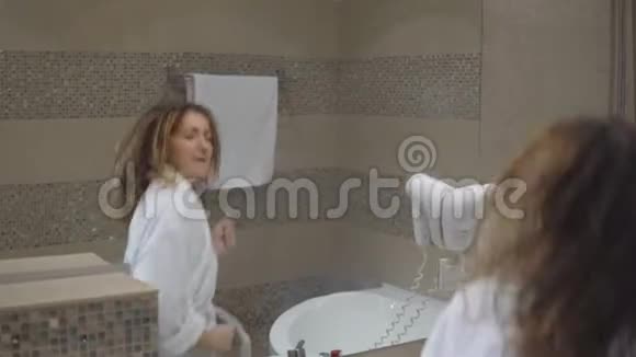 穿着白色长袍的有趣迷人的女孩洗了澡用吹风机在镜子前唱歌视频的预览图