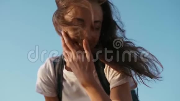 穿着白色t恤的年轻漂亮快乐的女人站在蓝天的背景上手挽直头发看着视频的预览图