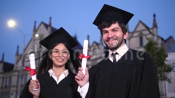 两个快乐的毕业学生摆着相机展示他们的文凭风把女孩的头吹掉了视频的预览图