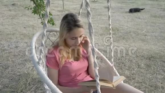 一个年轻的女学生在日落时在公园里读一本关于秋千的书她把头发伸直在白色上摆动视频的预览图