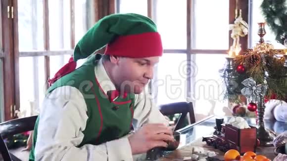饥饿的雄性精灵在圣诞节吃甜点穿着绿色服装的精灵在室内喝茶在圣诞老人旁边放着糕点视频的预览图