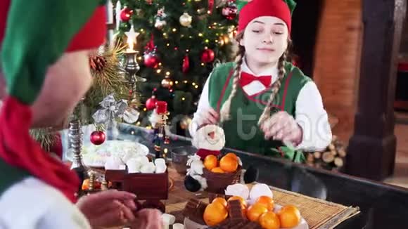 两个有趣的精灵坐在圣诞背景的桌子上女孩精灵试图带走糖果另一个精灵干扰了她视频的预览图