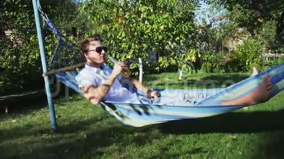 在花园的吊床上休息时冷静而又满足的迷人男人喝了一杯啤酒视频的预览图