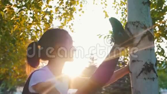 年轻迷人的男妈妈在公园里的树旁阳光下伸展抬起腿靠在树上视频的预览图