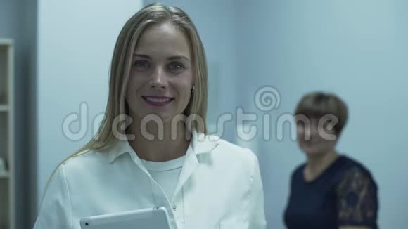 可爱的职业白种人医生微笑在前景看着相机而她成熟的病人坐在视频的预览图