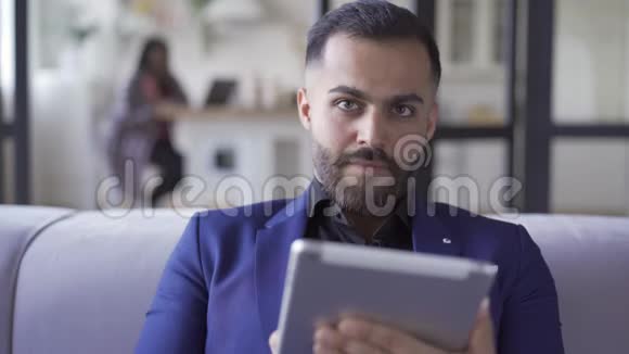 漂亮的长胡子男人穿着蓝色西装坐在家里用他的平板电脑工作室内休闲商人休息视频的预览图