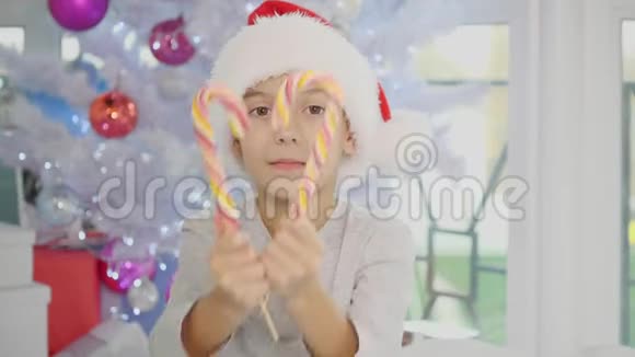 一个穿着圣诞帽的滑稽男孩穿着睡衣坐在床上向镜头伸出条纹糖果棒眨眼视频的预览图