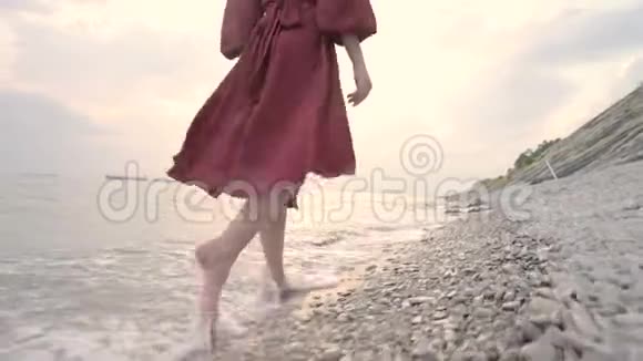 一个年轻女孩的腿穿着红色的裙子从后面走在岩石海滩上日落时海岸一件飘舞的衣服视频的预览图