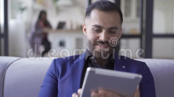 戴着蓝色西装的满脸胡须的男人坐在家里用他的平板电脑工作室内休闲商人休息后视频的预览图