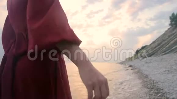 一个年轻女孩的手特写她穿着一件红色的裙子从身后走在日落时海边的岩石海滩上A视频的预览图