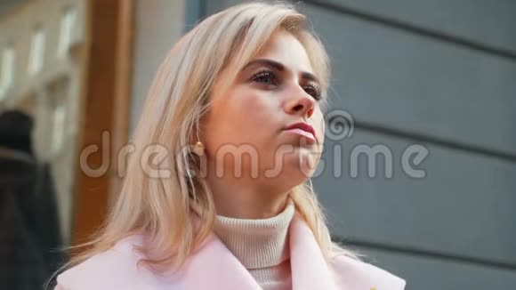 在购物中心的背景下站在街上的一位身穿粉红色外套的年轻漂亮金发女子的肖像画视频的预览图