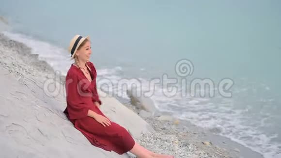 一个快乐的微笑女孩穿着红色的夏装和草帽坐在岩石海岸上看着大海的地平线他是视频的预览图