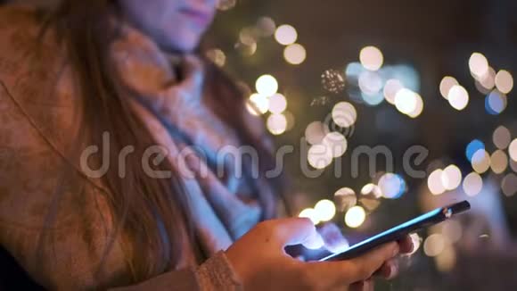 在冬天一个装饰着灯光的圣诞树的背景下女人的特写镜头使用智能手机通信视频的预览图