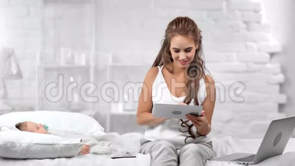 在小宝宝睡在白色卧室的时候妈妈用平板电脑多种多样的微笑聊天视频的预览图