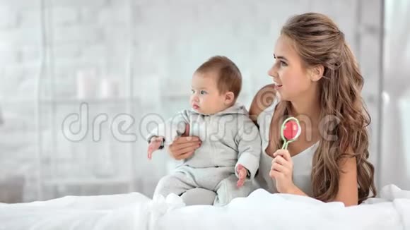 迷人的金发女人微笑着摇着拨浪鼓和小宝宝在白色卧室的内部休息视频的预览图