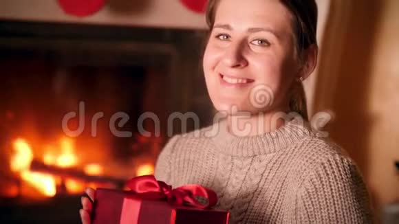 微笑的年轻女子在客厅里拿着红色礼品盒和燃烧的壁炉的画像视频的预览图
