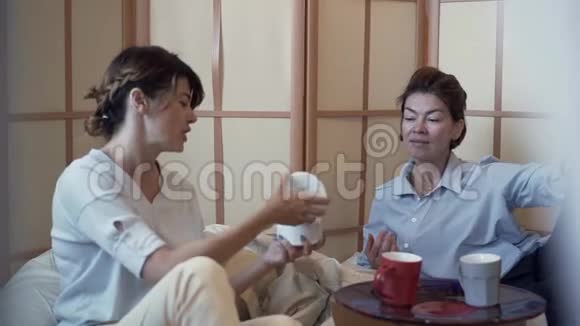 两个可爱的成熟女人坐在舒适的枕头椅上喝着咖啡谈论着视频的预览图