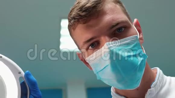 年轻英俊的牙医戴口罩在病人检查时戴着医疗灯并靠近视频的预览图