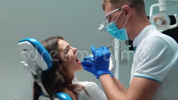 年轻的牙医戴着医用手套戴着口罩检查年轻的黑发女子病人使用牙科器械的牙齿问题视频的预览图
