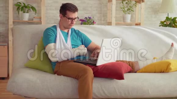 一个手臂和腿骨折的年轻人坐在沙发上用笔记本电脑的肖像视频的预览图