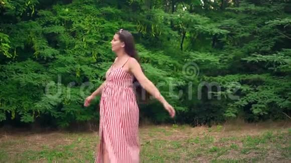 一个怀孕的女孩穿过公园一个穿着条纹白红色连衣裙的女孩微笑着在树间旋转视频的预览图