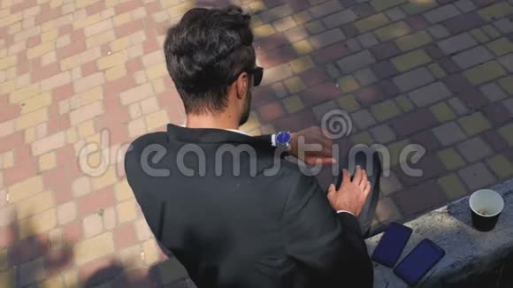 穿着经典西装和太阳镜的时髦男人坐在一个混凝土长凳上的公园里挺直了头发4k4k视频视频的预览图