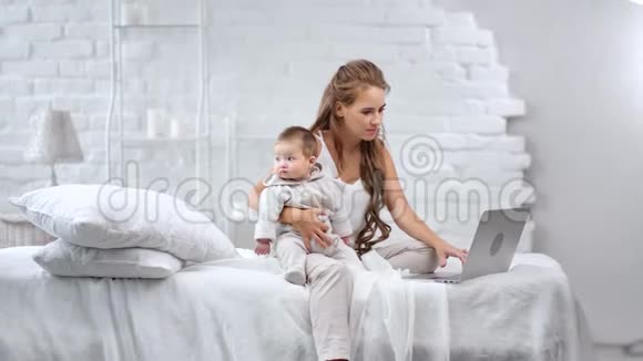 年轻的爱笑的妈妈多用笔记本电脑聊天抱着可爱的宝宝享受母性视频的预览图