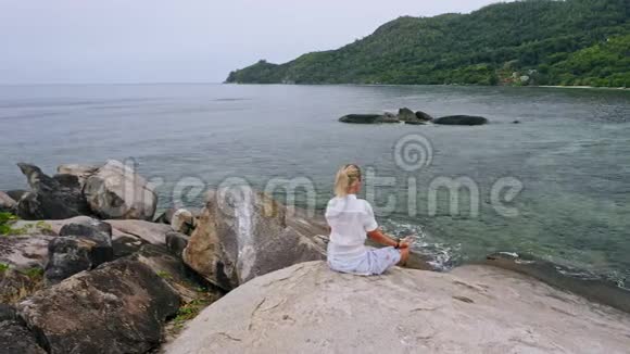 空中盘旋的镜头金发女孩坐在花岗岩岩石上的瑜伽姿势周围是异国情调的热带泻湖视频的预览图