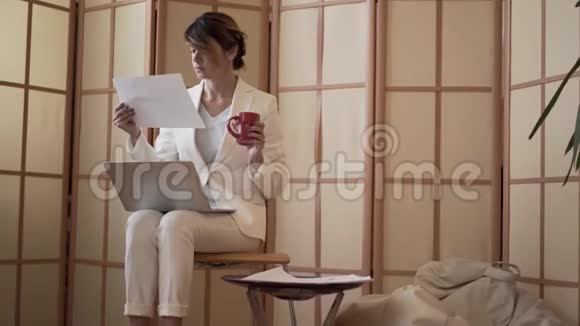 成熟的白种人商务女性穿着白色正式西装喝着咖啡看着笔记本电脑屏幕和纸坐着视频的预览图