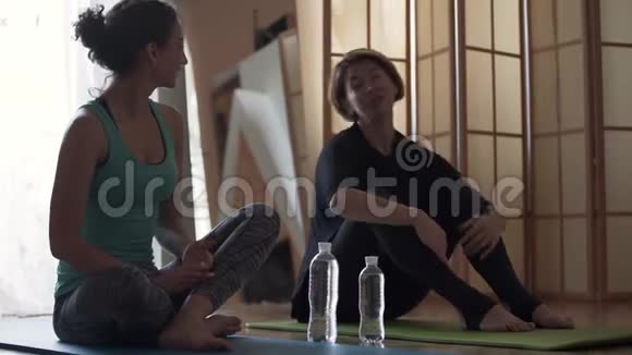 年轻女子带着私人教练坐在地板上的垫子在家里年长和年轻妇女伸展身体视频的预览图