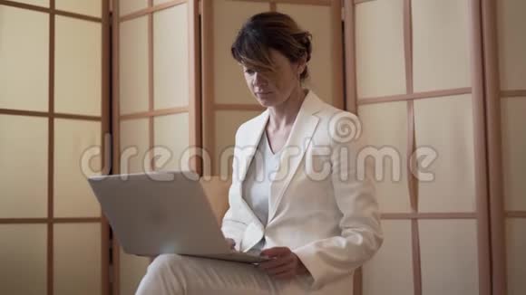 自信的女人穿着白色西装带着笔记本电脑坐在家里或现代办公室里工作女商人完成视频的预览图