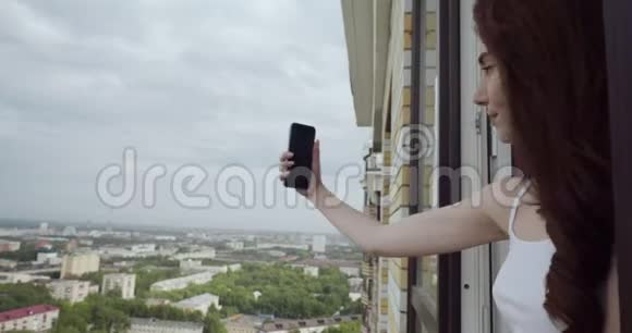 年轻女子从窗口看在智能手机上自拍视频的预览图