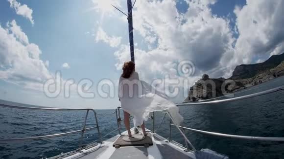 一个红头发的女孩站在一艘游艇的船头上它沿着岩石海岸沿着大海航行视频的预览图