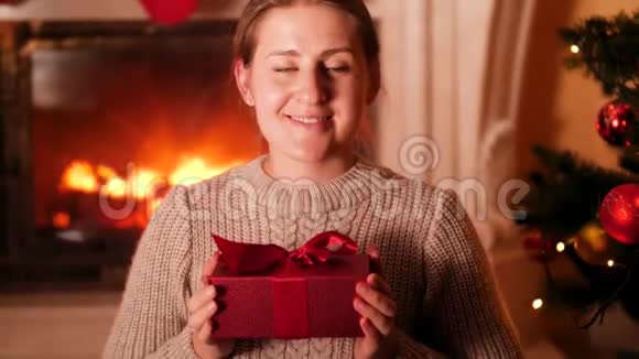 4k视频微笑的女人坐在燃烧的壁炉和赤柱沙树并向相机展示礼物盒视频的预览图
