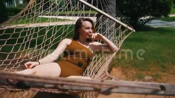 一位年轻漂亮的女士穿着一件泳衣躺在吊床上围着热带植物休息视频的预览图