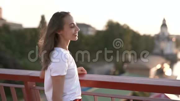 穿着白色t恤的年轻漂亮女人正站在城市背景上的桥上兴高采烈地展现在前方视频的预览图
