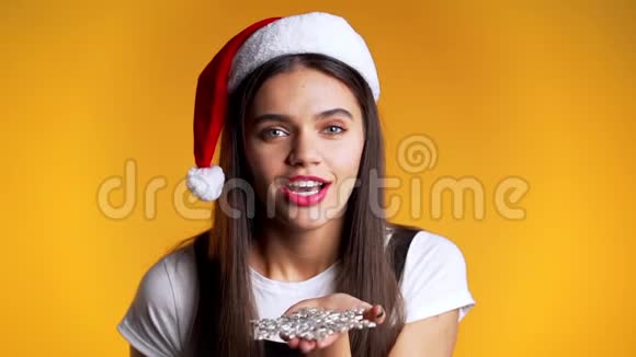 戴着圣诞帽的漂亮女孩吹着黄色背景的雪花纸屑视频的预览图
