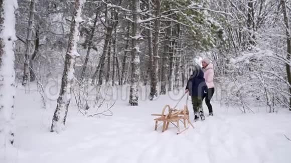 穿着温暖衣服的可爱男孩和他的母亲在下雪的冬日拉着木制的老式雪橇散步一个小男孩滑了进去视频的预览图