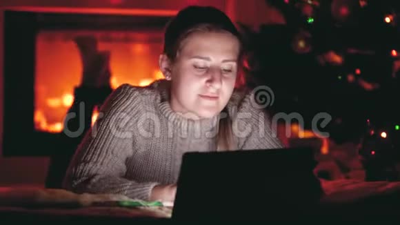 4k视频微笑的女孩在平板电脑上浏览互联网同时躺在壁炉旁和发光的圣诞树视频的预览图