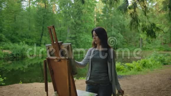 女艺术家穿着衬衫和牛仔裤在池塘附近的公园里画一幅风景画视频的预览图