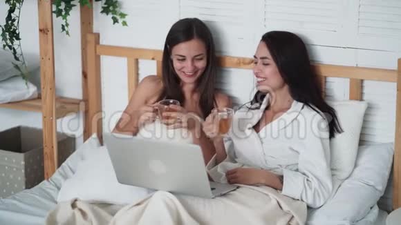 两个女朋友坐在床上喝茶在笔记本电脑上看有趣的电影笑视频的预览图