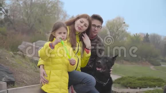 成年白种人妇女与大儿子小女儿和黑德伯曼一起坐在秋季公园家庭游戏视频的预览图