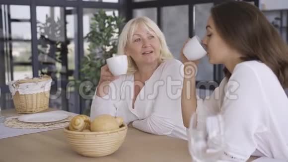 漂亮的成熟女人坐在厨房里女儿喝茶或咖啡分享她秘密的女孩视频的预览图