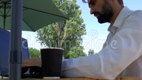 一个长着白衬衫胡子的帅哥坐在街边咖啡馆的桌子旁在笔记本电脑键盘上打字然后从一个视频的预览图