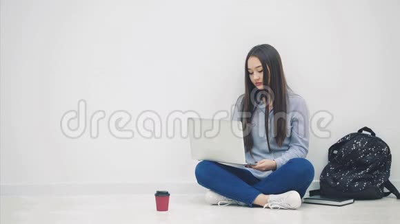 可爱的年轻亚洲女士坐在地板上以洛托斯的姿势在互联网上搜索挥手问好学校用品视频的预览图