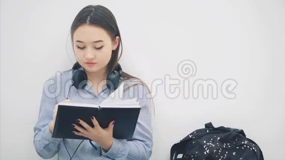 年轻的亚洲女孩坐在地板上厌倦了写作打哈欠所以她拿着手机和耳机听着视频的预览图