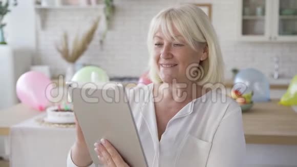 积极成熟的女人看着平板电脑微笑着站在现代厨房生日庆典视频的预览图
