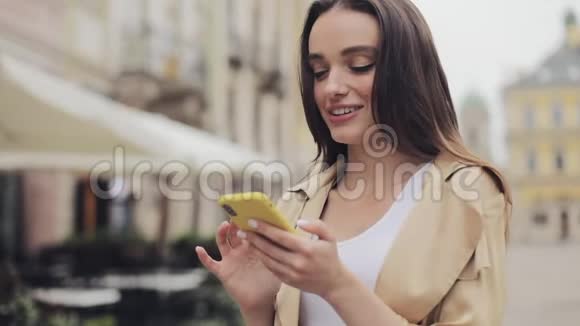 迷人的时尚年轻女孩使用她的智能手机打字和微笑站在老城市的背景特写视频的预览图