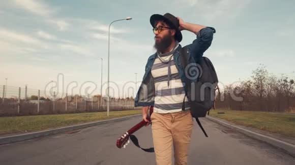 一位留着胡须的男音乐家在阳光明媚的日子里用一把吉他在公园里散步眼睛黝黑温暖视频的预览图